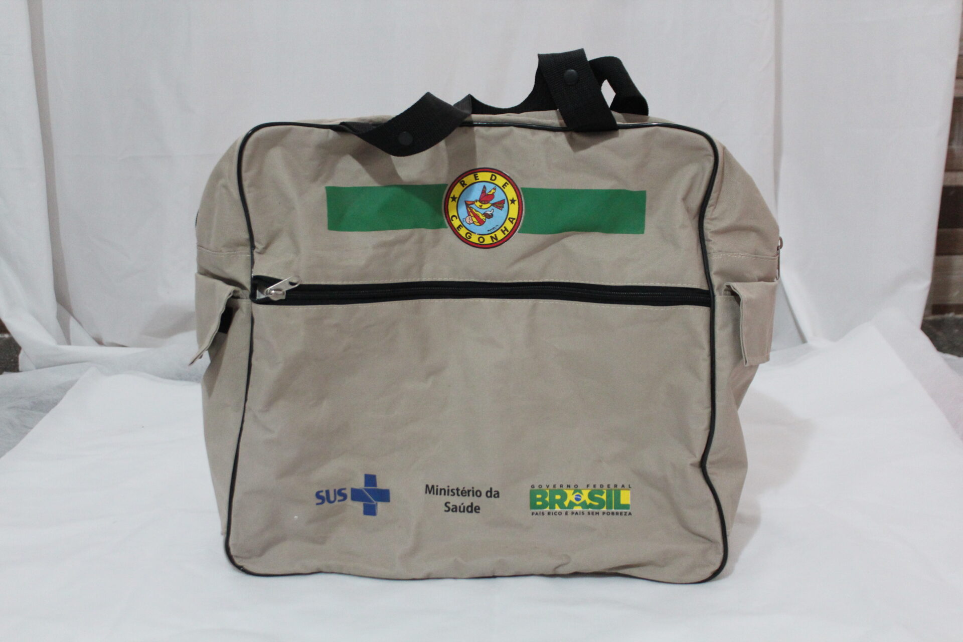 Bolsa de kit da parteira, entregue pela rede cegonha durante o governo Dilma, vista de frente. Tem formato retangular, bege, em tecido resistente, estilo poliéster, com as bordas contornadas com acabamento estreito e preto. Está apoiada sobre uma superfície clara, com fundo branco. 
A bolsa tem duas alças pretas, fixadas na parte central.
Na parte frontal, um bolso amplo com fechamento em zíper horizontal preto. 
Acima, uma faixa verde horizontal com um elemento circular central. Dentro dele, a ilustração de uma cegonha, em tons de vermelho e amarelo, com asas abertas, carregando um bebê envolto em pano vermelho, preso ao bico. Ao redor, há uma borda circular amarela contornada de vermelho, escrita em letras maiúsculas e pretas:  “REDE CEGONHA”.
Na parte inferior frontal, estão as logomarcas coloridas:  SUS, do Ministério da Saúde e do Governo Federal do Brasil,  “Brasil: País Rico é País sem Pobreza”.
