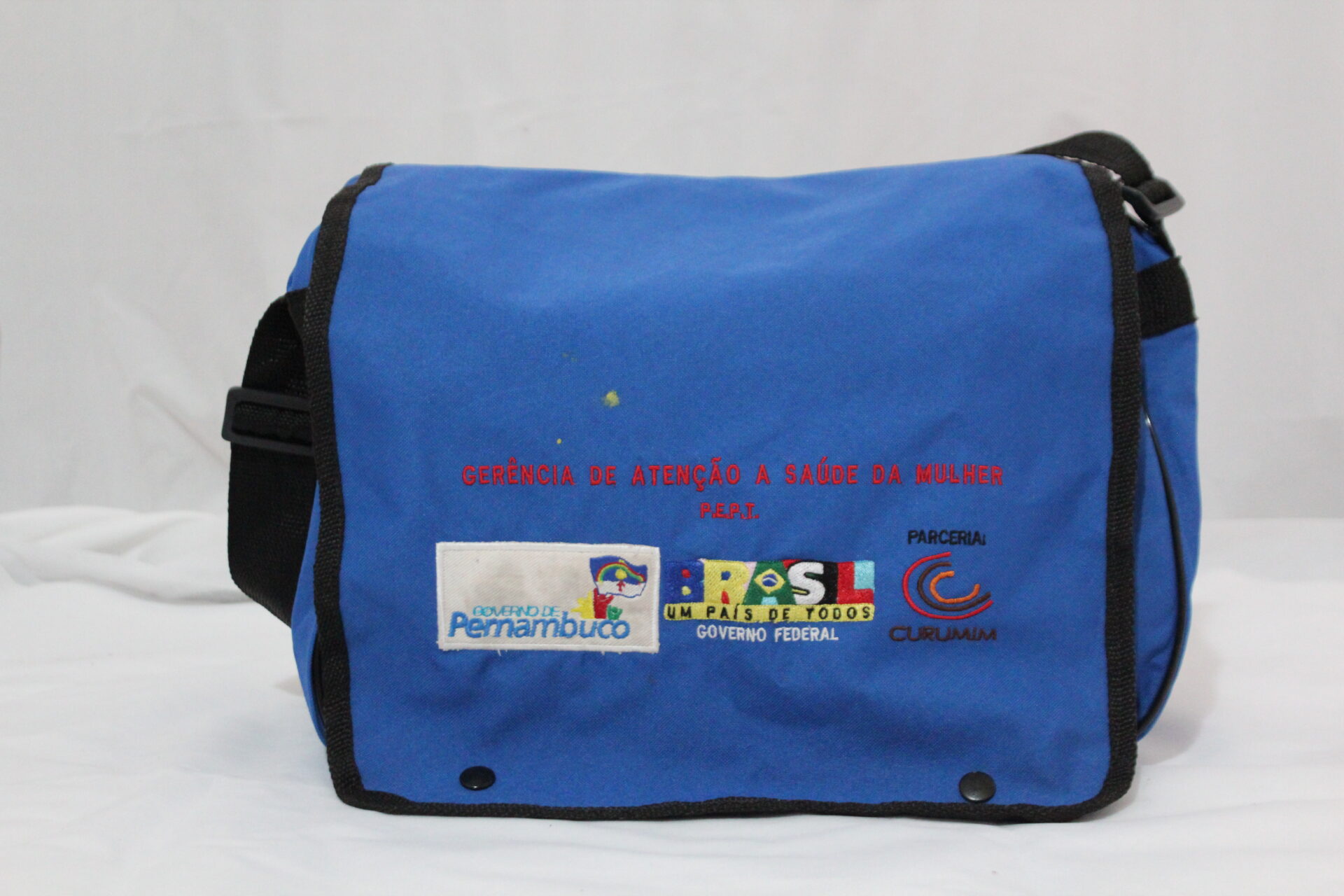 Bolsa de kit da parteira, Programa Estadual Parteiras Tradicionais (PEPT), vista de frente. Tem formato retangular horizontal, azul, em tecido resistente, poliéster, estilo carteira. Está apoiada sobre uma superfície clara, com fundo branco.
A bolsa tem aba frontal ampla, com fechamento por dois botões pretos de pressão na parte inferior e tem alça longa preta, ajustável, fixada nas laterais.
As bordas da aba da bolsa são contornadas por acabamento estreito preto.
Na parte frontal da aba tem em letras vermelhas: “GERÊNCIA DE ATENÇÃO À SAÚDE DA MULHER” e “P.E.P.T.” Abaixo, os logos: “Governo de Pernambuco”, “BRASIL:  Um País de Todos: Governo Federal” e  parceria “Curumim”.
