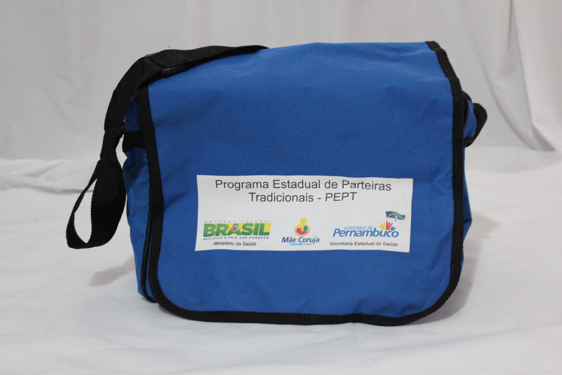 Bolsa de kit da parteira, Programa Estadual Parteiras Tradicionais (PEPT), vista de frente. Tem formato retangular horizontal, azul, em tecido resistente, poliéster, estilo carteira. Está apoiada sobre uma superfície clara, com fundo branco.
A bolsa tem aba frontal ampla sem fechamento aparente e alça longa preta, ajustável, fixada nas laterais. As bordas da aba são contornadas por acabamento estreito em preto.
Ao centro da parte da frente da bolsa, há um retângulo branco que tem em preto na parte superior, a informação: “Programa Estadual de Parteiras Tradicionais (PEPT) e abaixo, os logos coloridos: “BRASIL País Rico é País sem Pobreza, Ministério da Saúde”, “Mãe Coruja Pernambuco” e “Governo de Pernambuco: Secretaria Estadual de Saúde”.