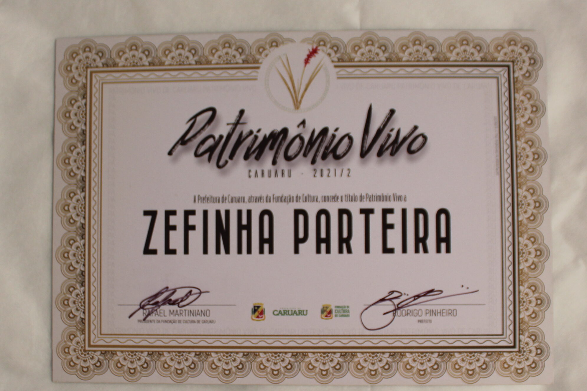 Certificado de título de patrimônio vivo concedido a Zefinha Parteira. Certificado na horizontal,  em papel branco, contornado por uma moldura decorativa dourada que lembra flores em arabescos.
No topo, centralizado, uma plantinha estilizada com folhas alongadas douradas e uma flor em formato de espiga alongada vermelha.
Abaixo, o título em fonte cursiva e preta: “Patrimônio Vivo”. O nome da homenageada Zefinha Parteira está em destaque com letras grandes e de imprensa.
Informações textuais: 
Patrimônio Vivo
CARUARU - 2021/2
A Prefeitura de Caruaru, através da Fundação de Cultura, concede o título de Patrimônio Vivo a ZEFINHA PARTEIRA
Na parte inferior:
As assinaturas e os nomes:  RAFAEL MARTINIANO, Presidente da Fundação de Cultura e RODRIGO PINHEIRO, Prefeito.
Entre as assinaturas, estão os logos: Prefeitura de Caruaru e da Fundação de Cultura.
