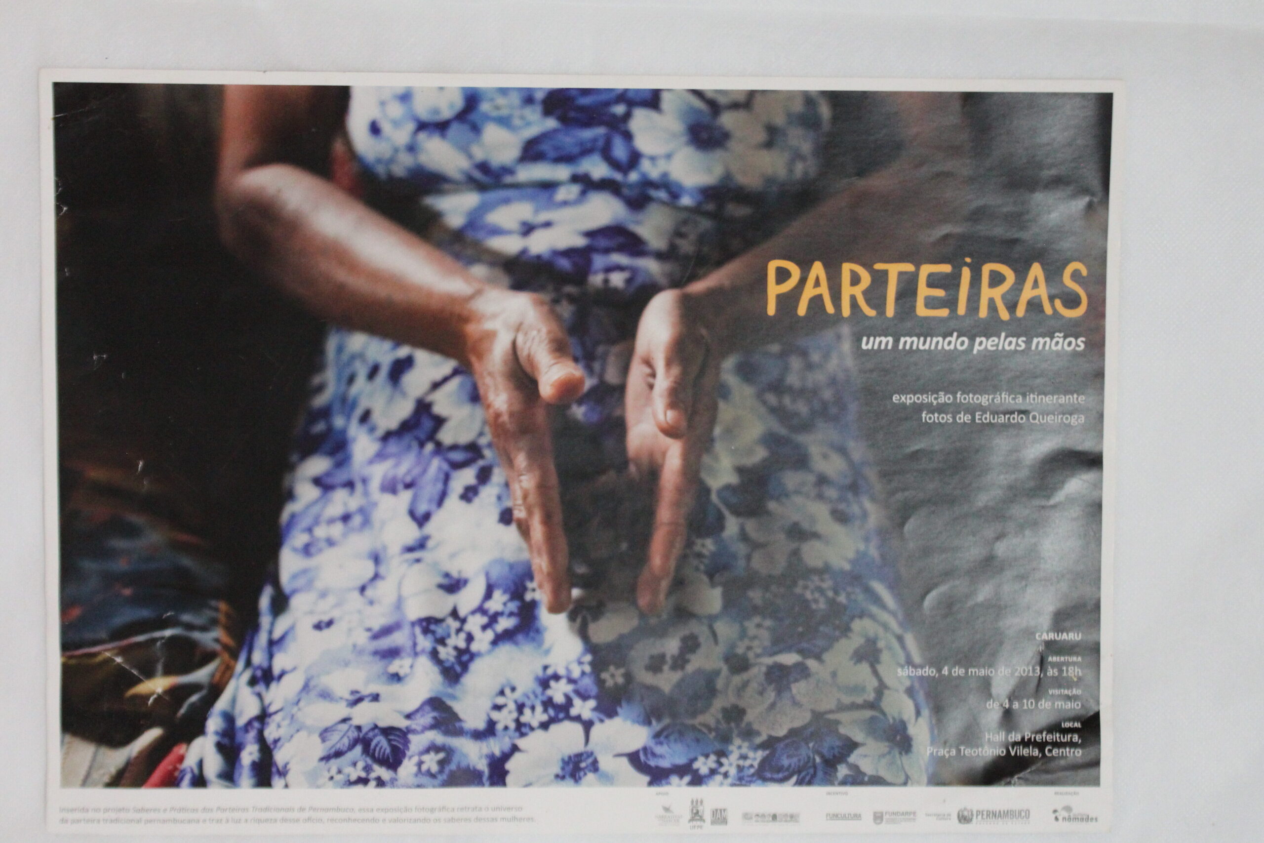 Cartaz colorido, em formato horizontal, com fotografia ocupando toda a extensão da imagem, uma mulher negra é vista do busto até a altura dos joelhos.  Ela está com as palmas das mãos voltadas para dentro, levemente afastadas, e direcionadas para baixo, posicionadas à frente do ventre. A pele negra recebe uma luz natural que a faz brilhar. A mulher usa vestido florido em tons suaves de azul e branco. Atrás dela, o ambiente é escuro e desfocado.
À direita da mulher, sobre a área escura, em amarelo a palavra: “PARTEIRAS”, em letras grandes. Abaixo, em letras menores: “um mundo pelas mãos”.“Exposição fotográfica itinerante” e “fotos de Eduardo Queiroga”.
Próximo à margem inferior, à direita, as informações do evento: “Caruaru”, “abertura sábado, 4 de maio de 2013, às 16h”, “visitação de 4 a 10 de maio”, “local Hall da Prefeitura, Praça Teotônio Vilela, Centro”.
Na margem inferior, há uma faixa estreita e branca com logotipos institucionais borrados.